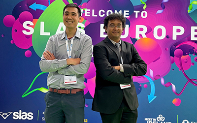 SLAS Europe 2022 Student Poster Award winners Winston Chiu, Ph.D. and Srijit Seal, Ph.D. standing in front of the SLAS 2022 Europe Logo.